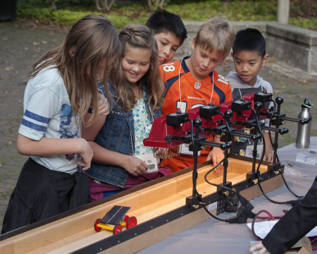 A group of 5 students watch their small solar powered car race down a wooden track.