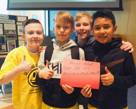 4 Lacamas Lake Elementary School Green Team members smile at the camera while holding a red paper sign with the handwritten words Any Age Can Be An Environmentalist