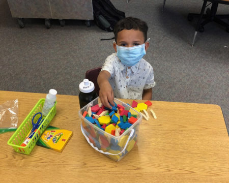 Kindergarten student sits at a large table wearing a mask.