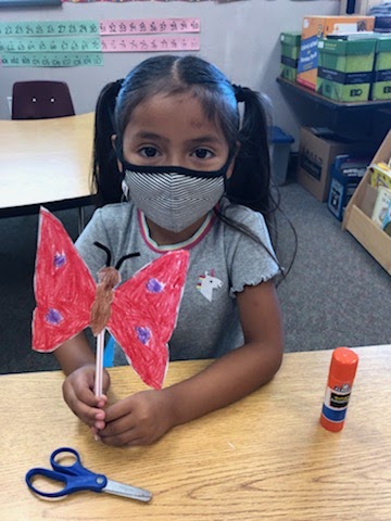 A young girl wears a mask while holding a homemade pink paper butterfly