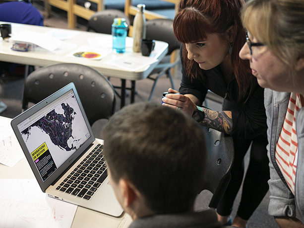 Three teachers are looking at a laptop's computer screen that displays a map of North America with migration patterns.