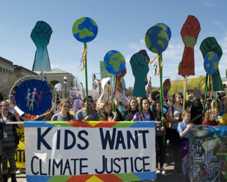 Group of kids at protest all behind a large banner sign that reads Kids Want Climate Justice. There are many other large signs in the air of the earth and fists.