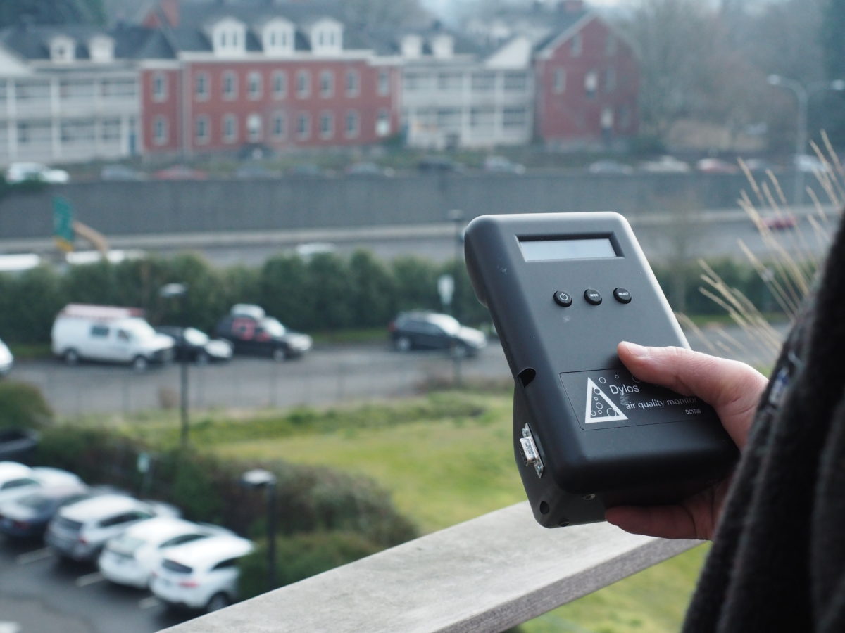 A close up image of an air quality meter in a student's hand overlooking a road and buildings