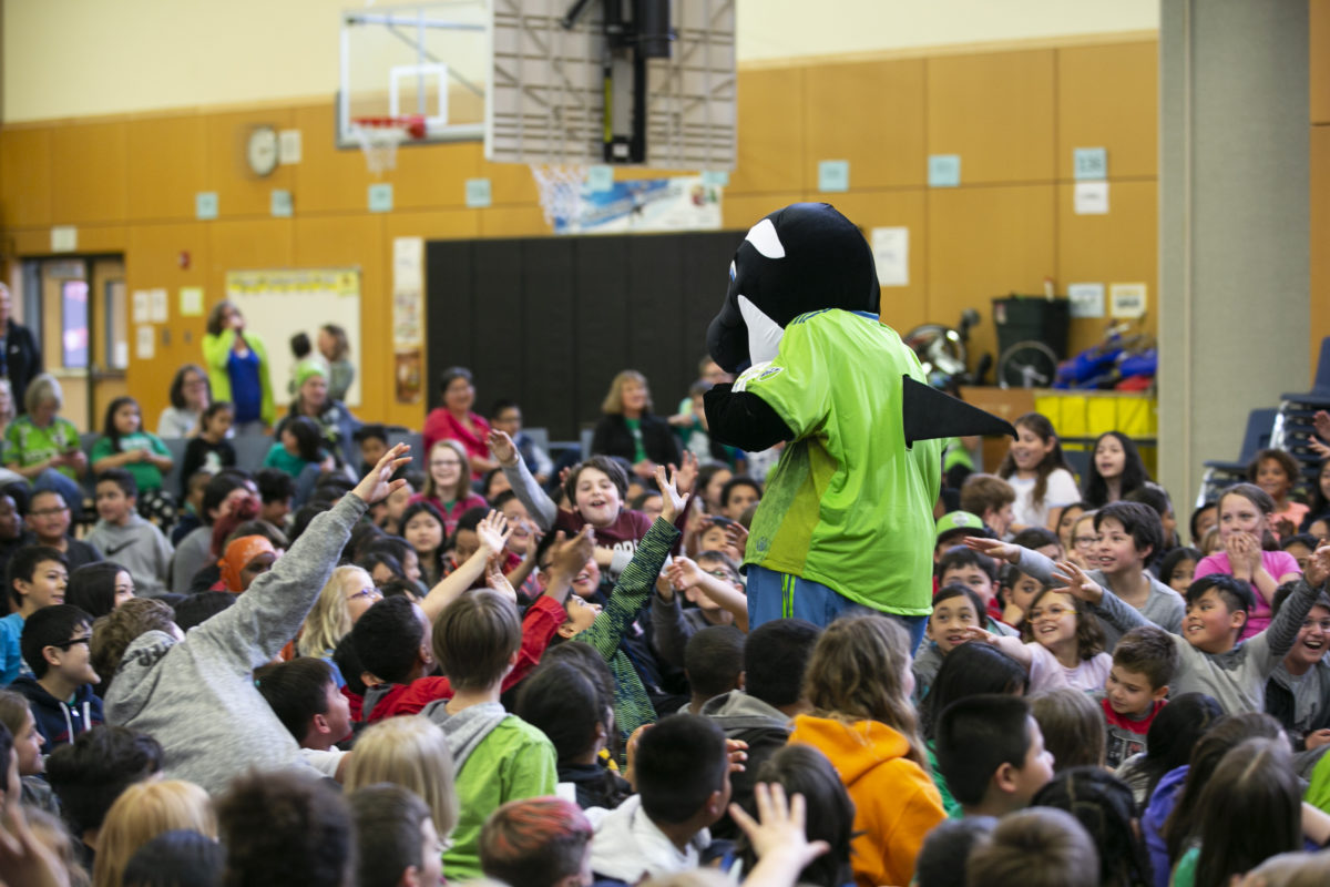 Sammy the Sounder stands surrounded by a large group of sitting children at a school assembly