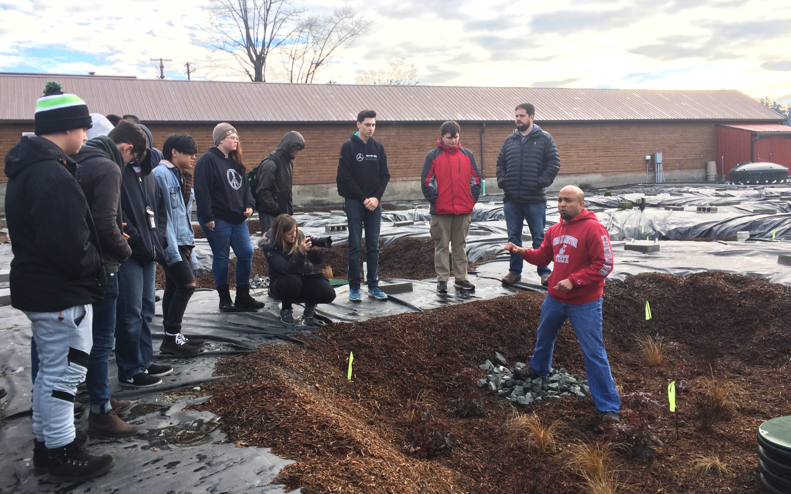 A group of students stands on the edge of an under construction rain garden listening to a rain garden expert talk about stormwater