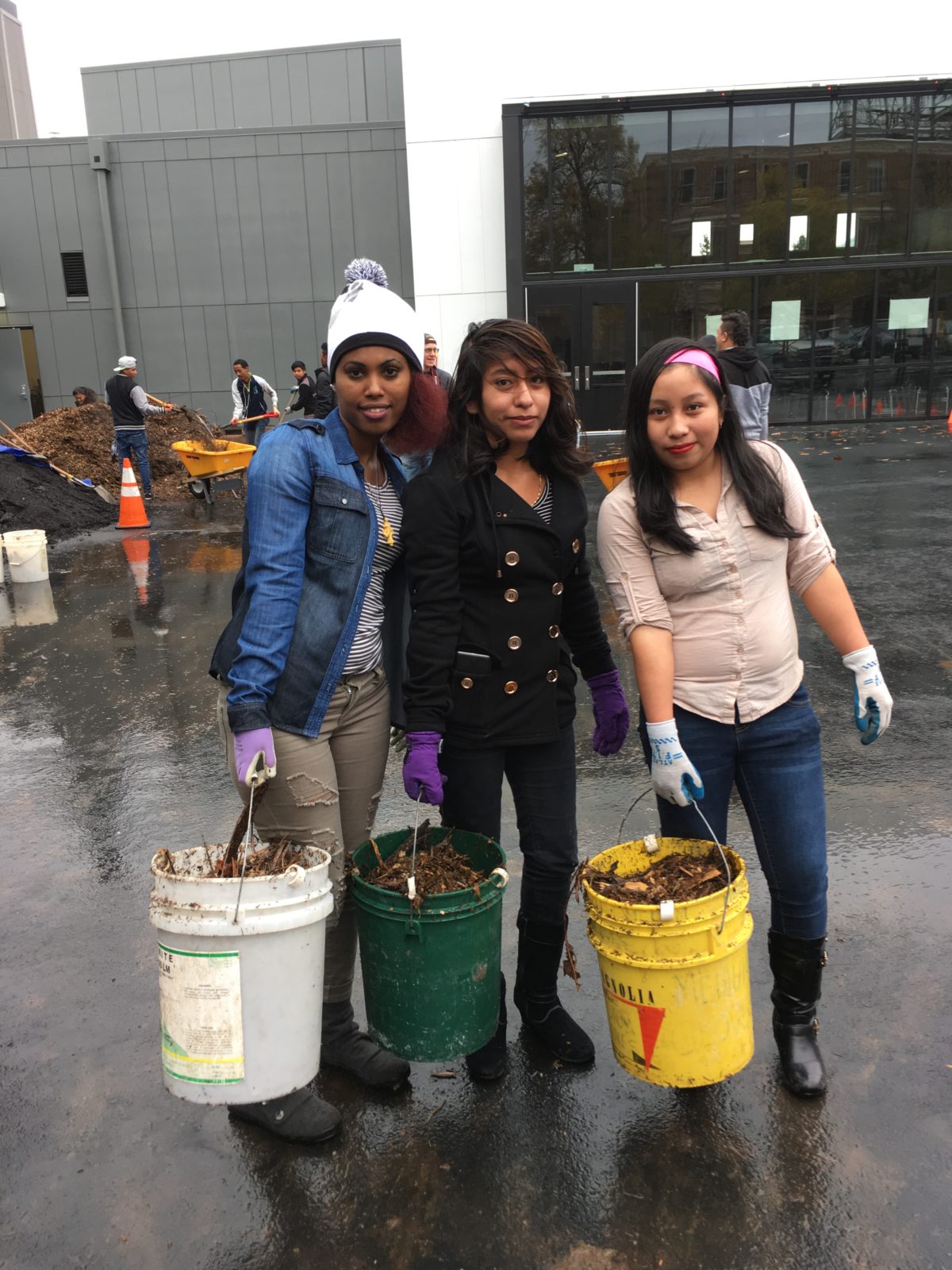 3 high school students hold buckets of mulch and smile at the camera