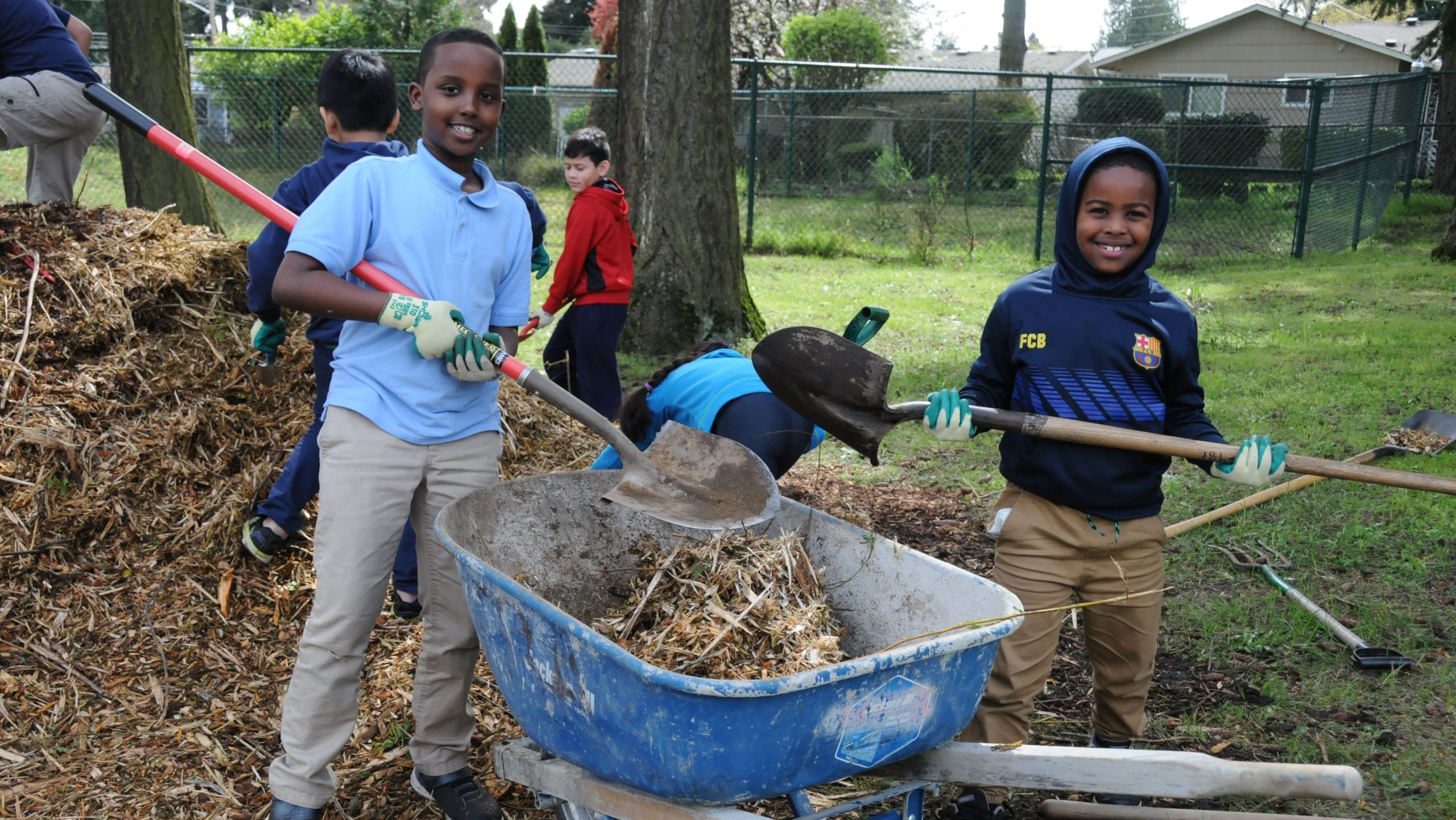 2 elementary school students pose for a picture holding shovels in front of a wheelbarrow full of mulch.