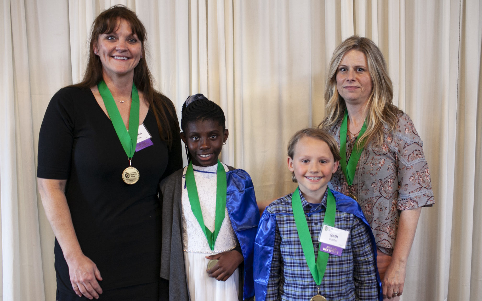 A group of 2 students and 2 teachers stands smiling at the camera with green medals around their necks.