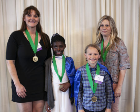 A group of 2 students and 2 teachers stands smiling at the camera with green medals around their necks.