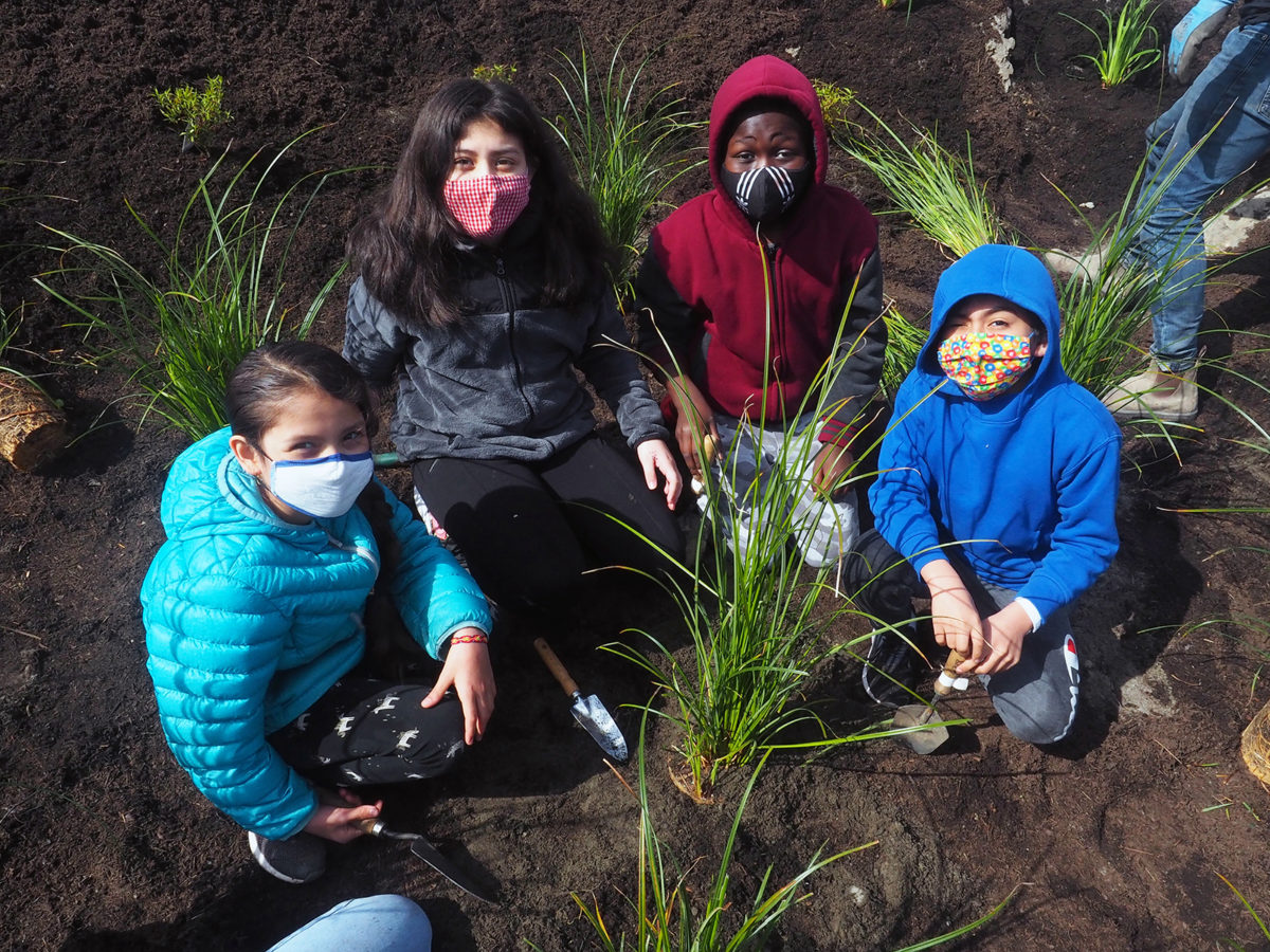 4 children looking at the camera pausing from planting plants in a new rain garden