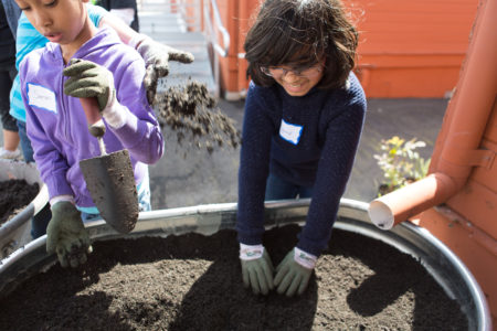 Youth digs in a raised planter bed while a pair of hands sprinkles soil near by.
