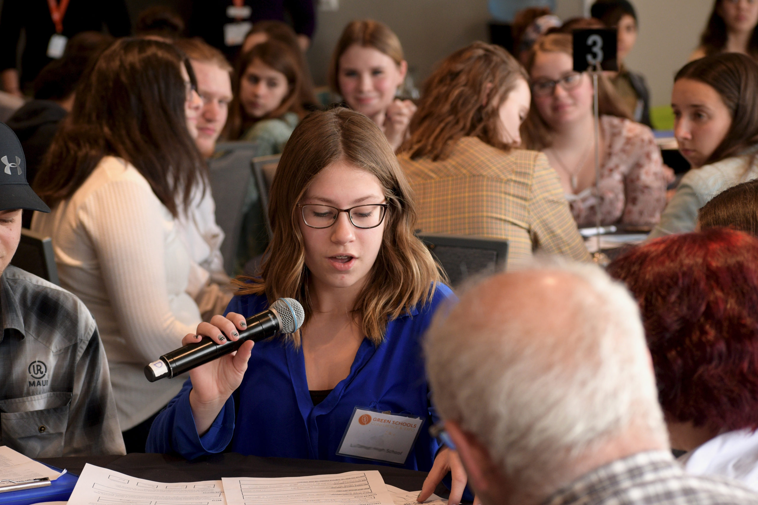 A student shares her thoughts at a previous Student Summit. She is holding a microphone and surrounded by other students.