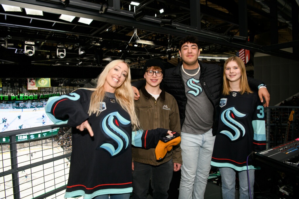 Silas High School teacher Breanne Willard and her three studetns wear Kraken jerseys and stand with the Seattle Kraken's rink behind them. 