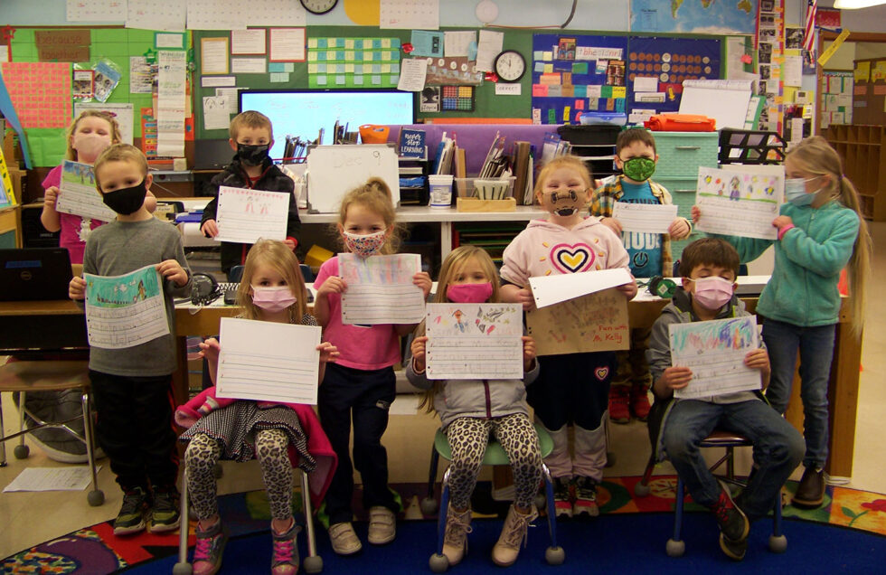 A group of 10 kindergarteners wears masks and looks at the camera holding up classwork as part of their marine debris lesson.