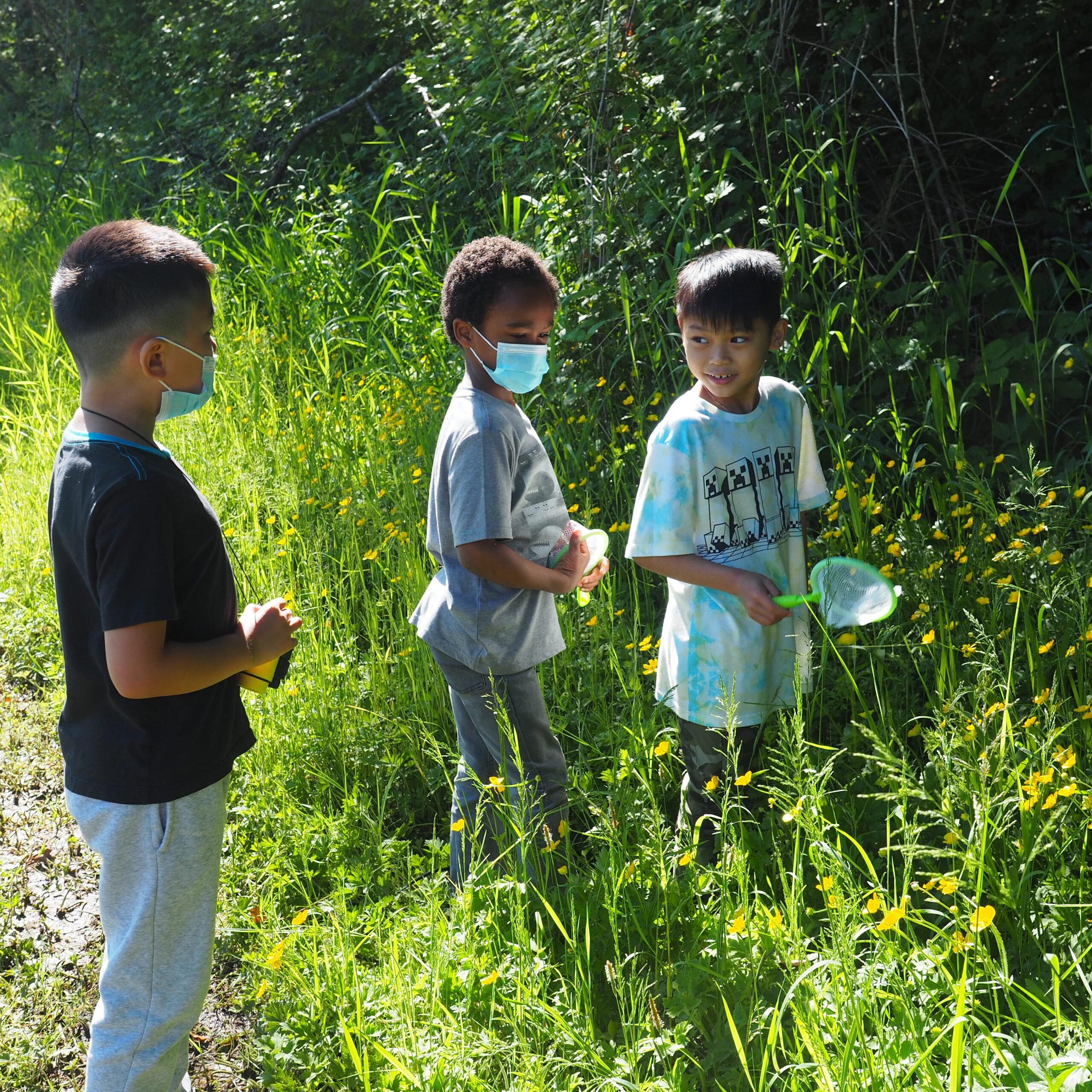 3 students stand in grass near their playground looking for pollinators