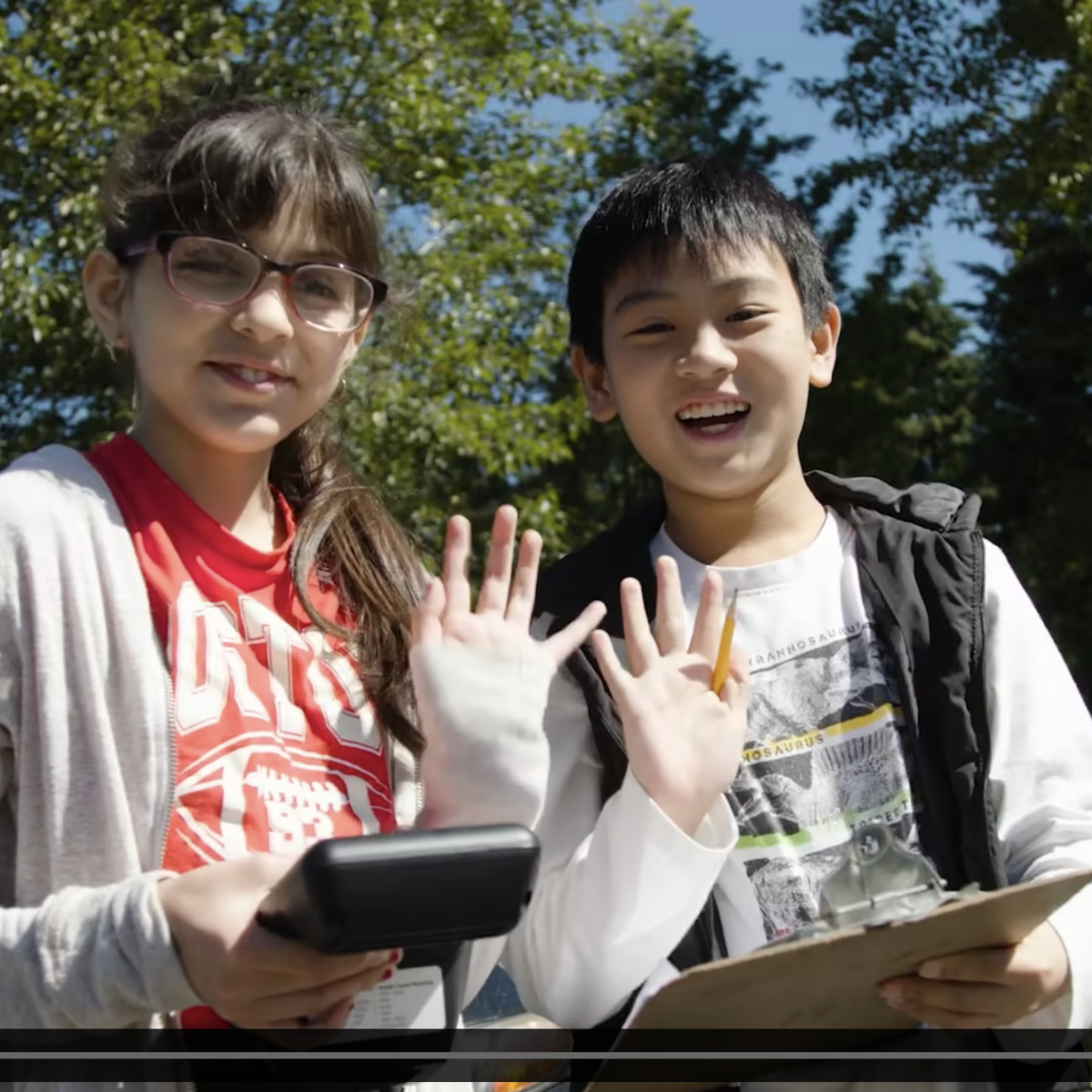 2 students look up to smile and wave at the camera. One is holding an air quality meter and the other is holding a clipbaord.