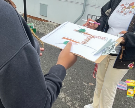 A barren brown patch of dirt and asphalt is visible between 2 portable buildings at Tyee's interim campus. Students hold a clipboard that shows their ideas to improve their campus.