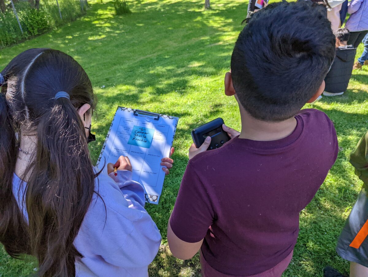 2 students look at an air quality monitor and write notes on a clipboard