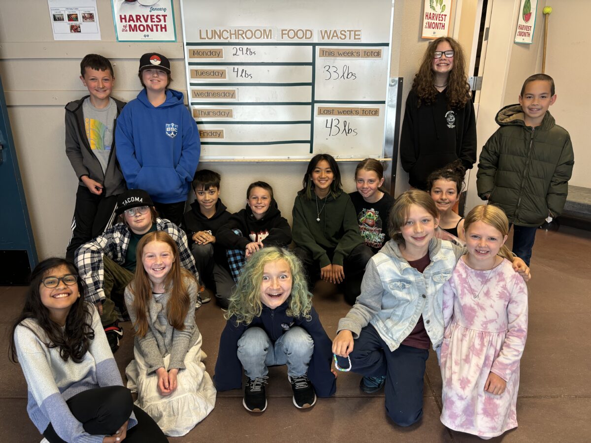 A group shot of students standing in front of a white board that tracks the food waste collected in their cafeteria.