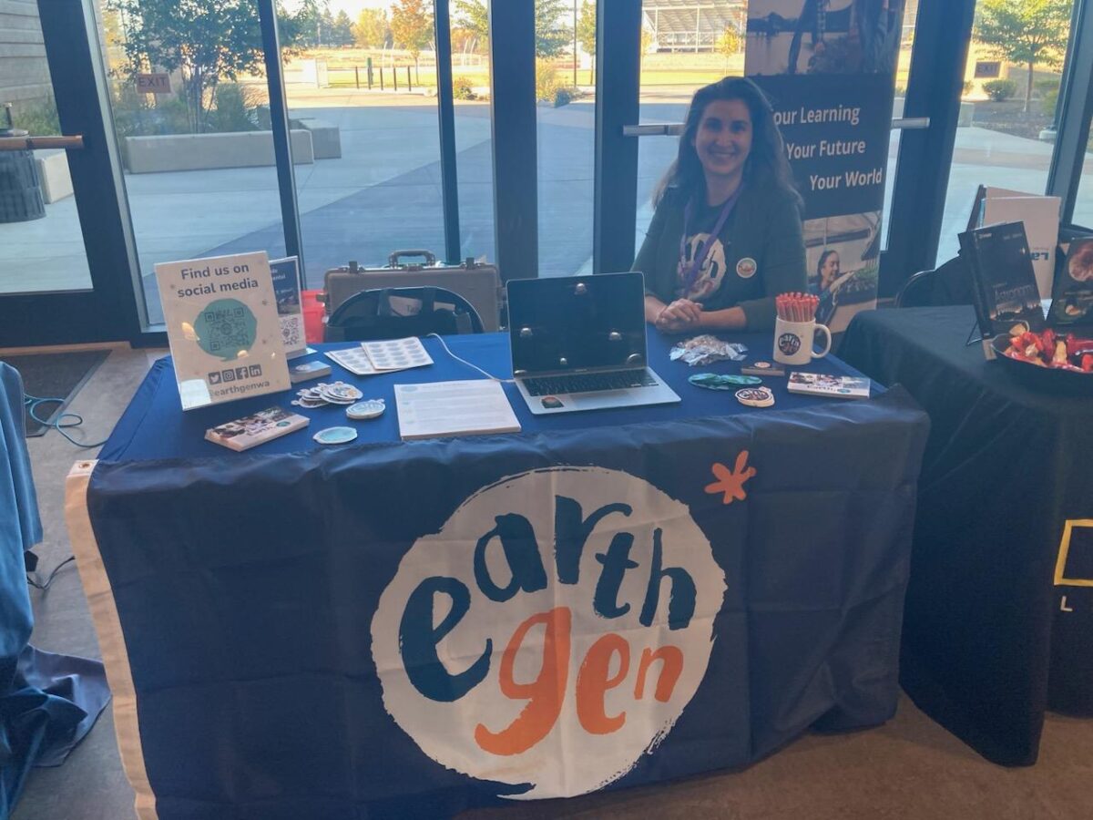  A photo of an EarthGen booth at an event. The table is covered with an EarthGen-branded tablecloth and features promotional materials, such as stickers, brochures, and a "Find us on social media" sign with EarthGen's handle. A staff member sits behind the table, smiling at the camera.