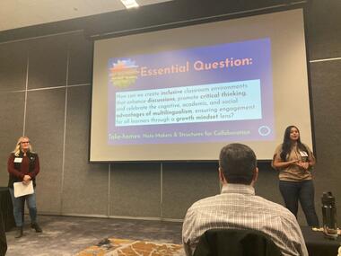 A photo of two presenters standing in front of a screen during a conference presentation. The screen displays an essential question for educators about creating inclusive classrooms that encourage critical thinking, academic growth, and a growth mindset through multilingualism and diversity.