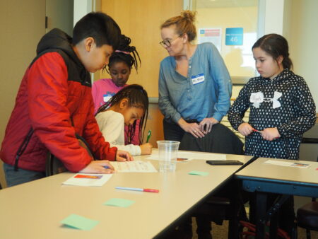 Four students collaborate on a group activity at a table while a teacher observes and offers guidance.