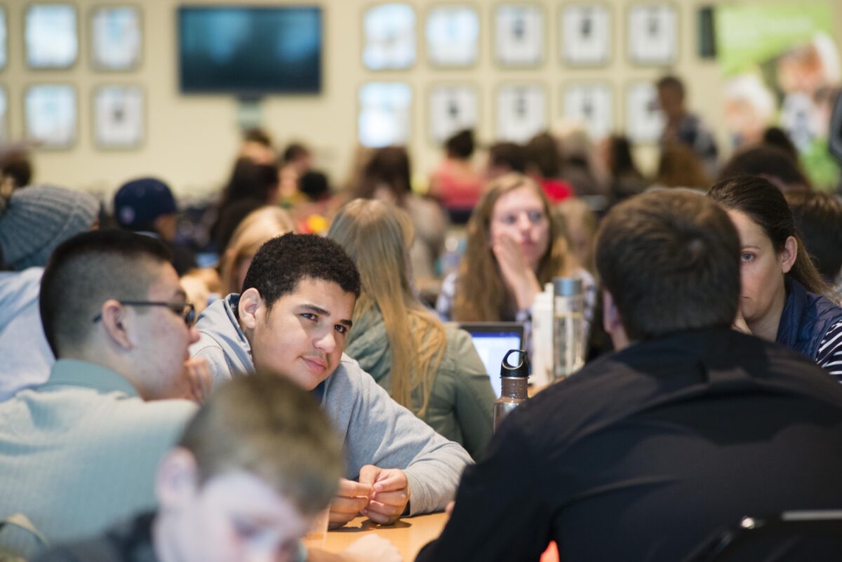 A large group of students gathered indoors, sitting at tables in conversation. A student in the center leans forward, listening and smiling during a discussion.
