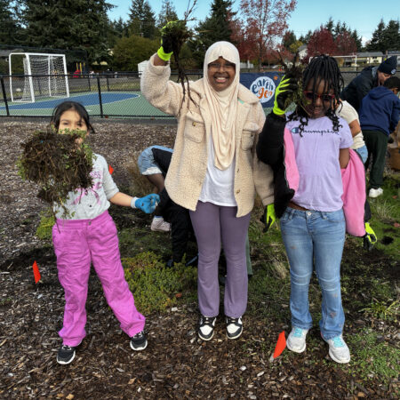 3 students hold up weeds they've pulled and they smile at the camera